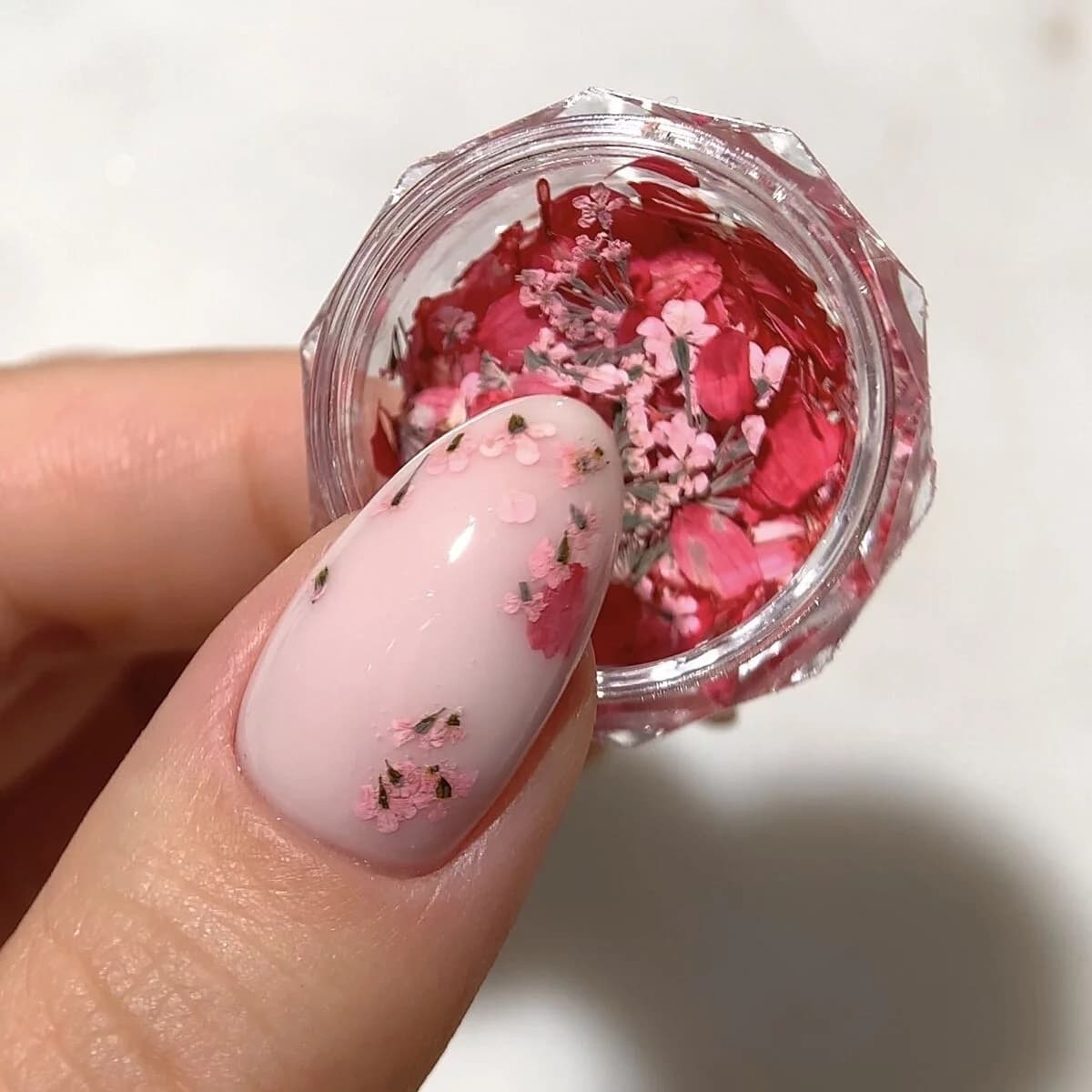 Un pot rempli de pétales de fleurs séchées dans des nuances rose corail - Semilac Flower Flakes Coral Blossom. Un ongle avec un vsp rose tendre est décoré de quelques pétales.