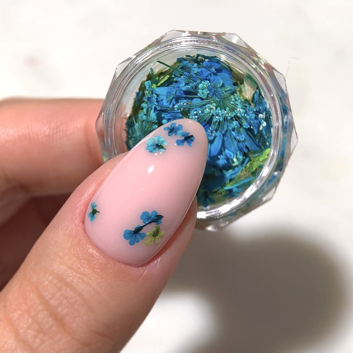 Un pot rempli de pétales de fleurs séchées dans des nuances pastel vert et turquoise - Semilac Flower Flakes Spring Meadow. Un ongle avec un vsp rose tendre est décoré de quelques pétales.
