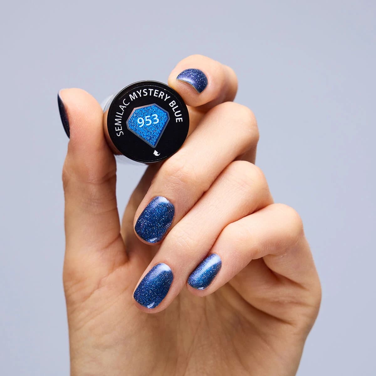 Main qui tient le flacon de Vernis semi-permanent 953 Mystery Blue de Semilac. Sur les ongles un bleu marine profond aux reflets irisés.